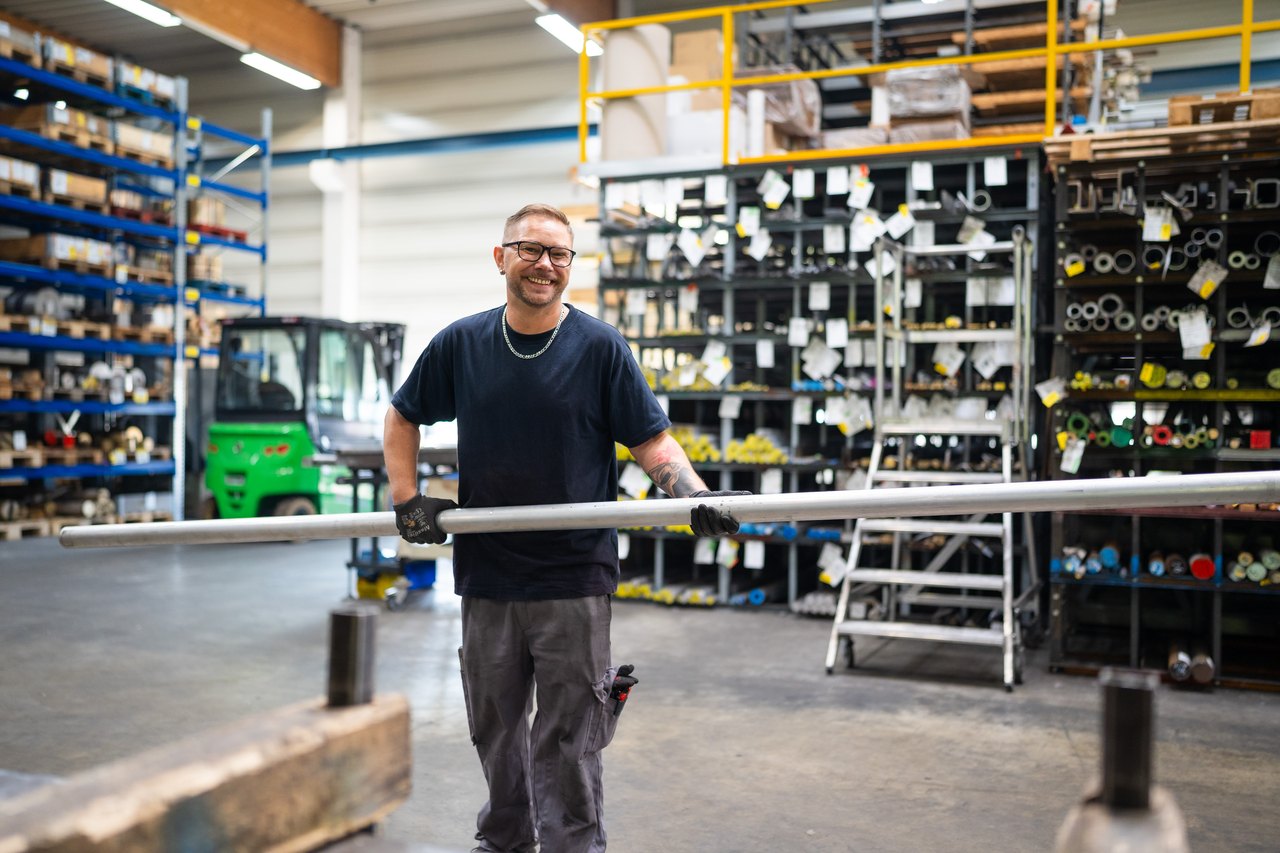 Ein Mann in Brille und Arbeitskleidung trägt zwei lange Metallrohre in einem Lagerhaus, umgeben von Regalen.